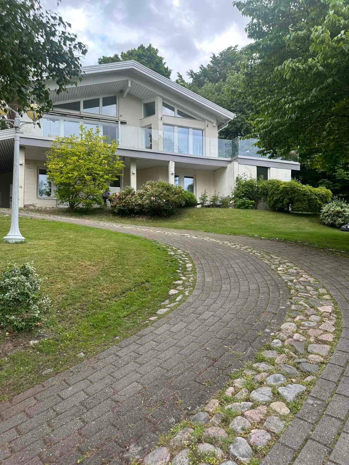 The exterior of a modern villa features a curved stone pathway leading to the entrance. Surrounded by well-maintained greenery and shrubs, the two-story structure includes large glass doors and a spacious balcony with railing. A lamp post adds a welcoming touch to the landscaped setting.
