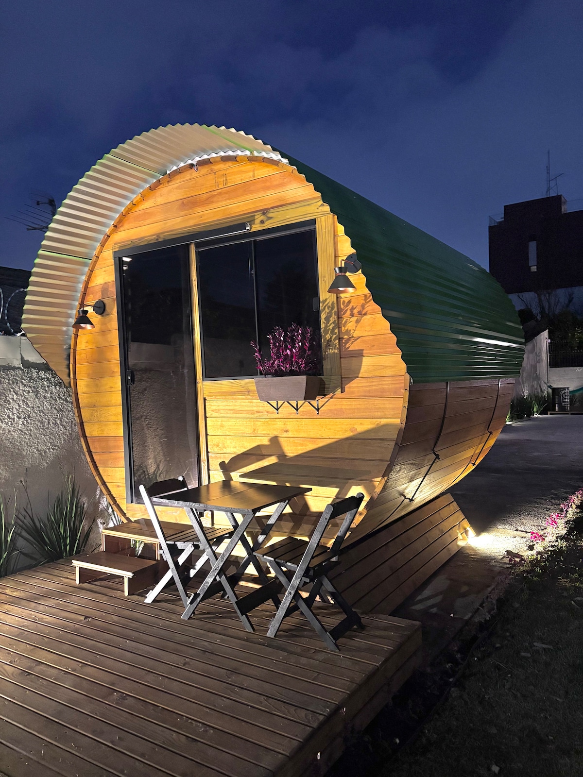 A uniquely designed round cabin features a wooden exterior with a green metal roof. Two foldable chairs are positioned on a wooden deck, with soft lighting illuminating the structure at dusk. A small flower box enhances the entrance, creating a welcoming atmosphere.