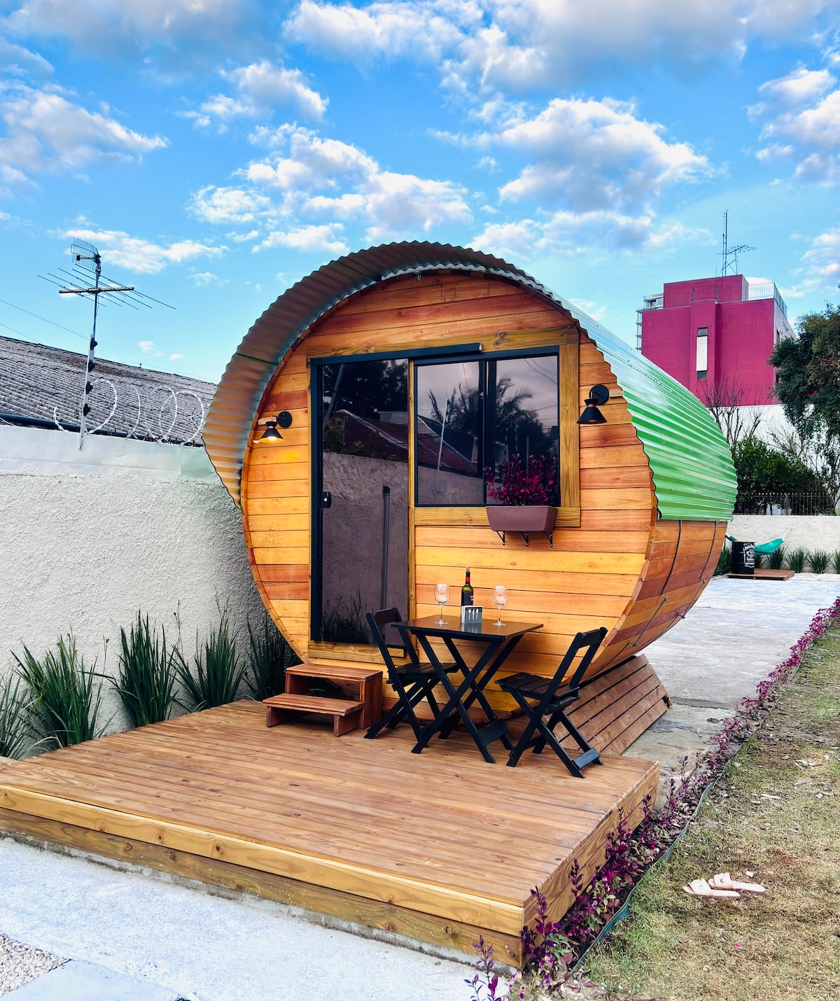 A unique architectural structure resembling a barrel, crafted from wood and featuring a green metal roof. A small wooden deck holds a table and two chairs, inviting relaxation. The surrounding area is landscaped with grass and plants, under a blue sky filled with scattered clouds.