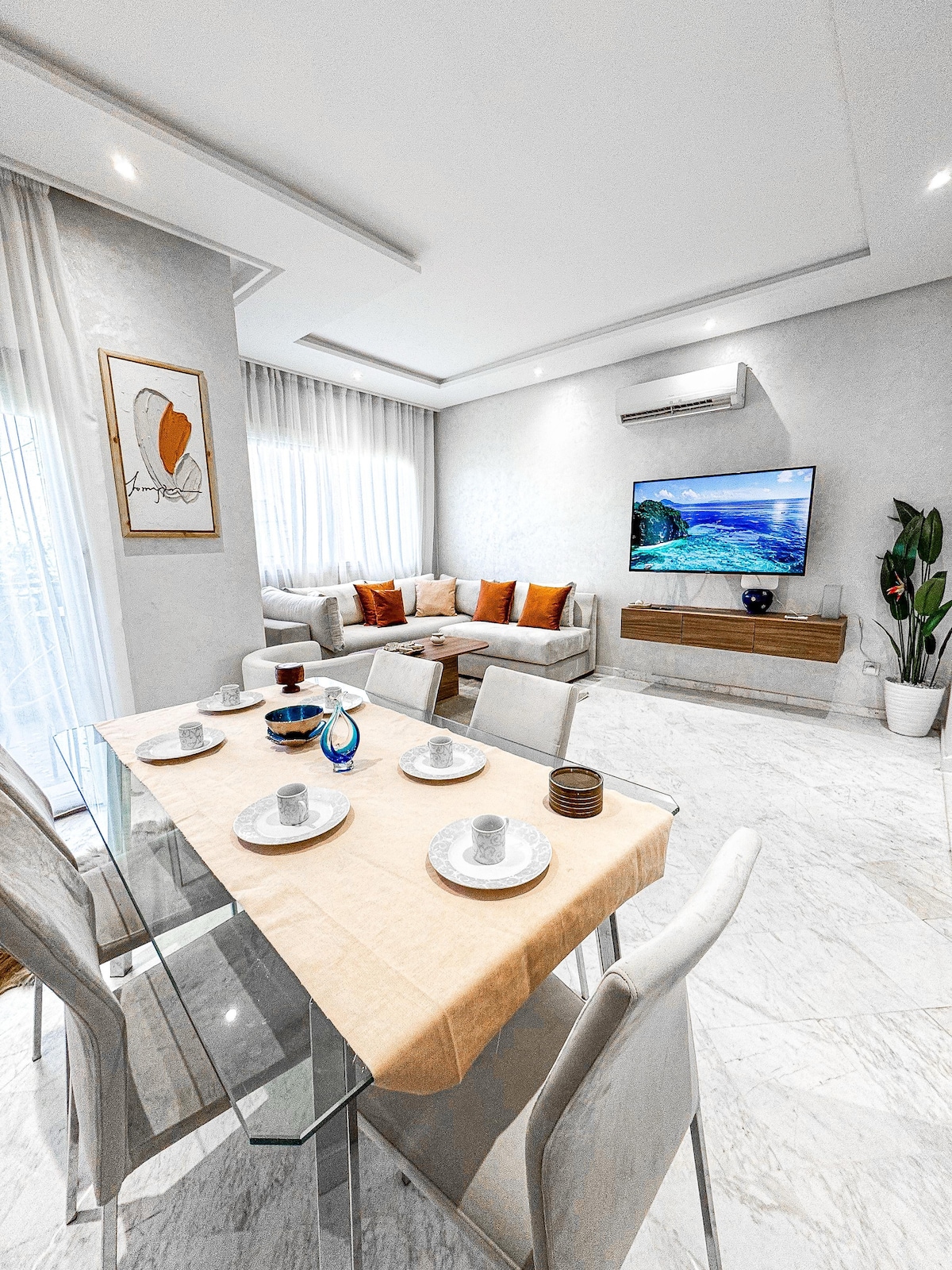 A modern dining area features a glass table set with plates and cups. Light-colored chairs surround the table. A cozy living space with a soft sofa and colorful cushions is visible in the background, alongside a television mounted on the wall, and a decorative plant.