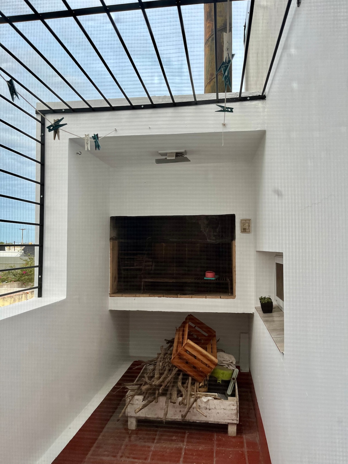 An outdoor space is enclosed by a grid structure, featuring a large wall opening that reveals a cozy area with gathered timber and a small potted plant. The floor is tiled in terracotta, contributing warmth to the minimalist design.