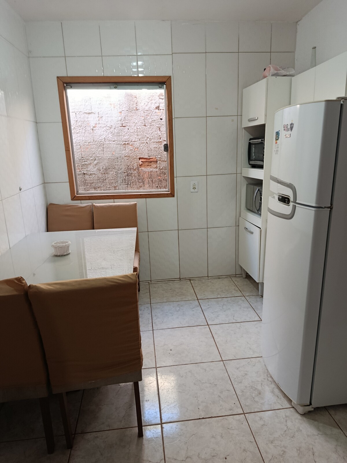 A compact kitchen showcases white tiled walls and a fitted dining table surrounded by cushioned chairs. A window allows natural light to brighten the space. A refrigerator and appliances are neatly arranged along one wall, contributing to a functional and organized environment.