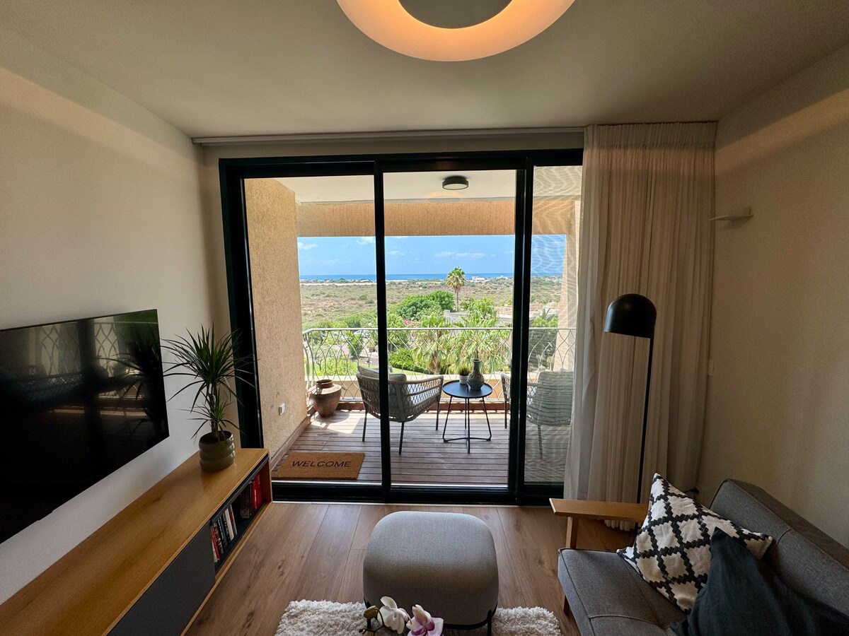 A spacious living area features a television mounted on a wooden console and a round ottoman. Large glass doors open to a balcony with seating, revealing a view of greenery and the sea in the distance, enhancing the airy ambiance of the space.