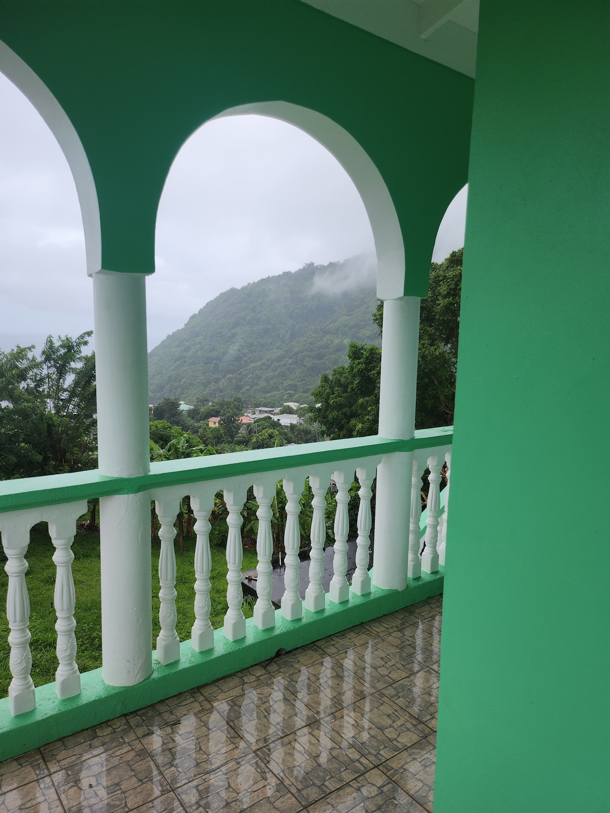 A balcony with green columns overlooks a lush hillside shrouded in mist. The area features white balustrades and tiled flooring, reflecting the rain. A vibrant green wall adds to the inviting aesthetic, while a serene landscape is visible in the distance.