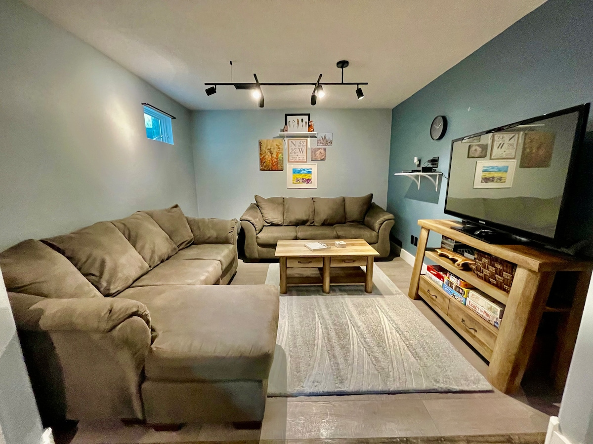 The living room features a spacious layout with a sectional sofa and a separate couch, offering ample seating. A flat-screen TV is positioned on a wooden console, complemented by a coffee table and light-colored area rug. Natural light enters through a small window, enhancing the inviting ambiance.