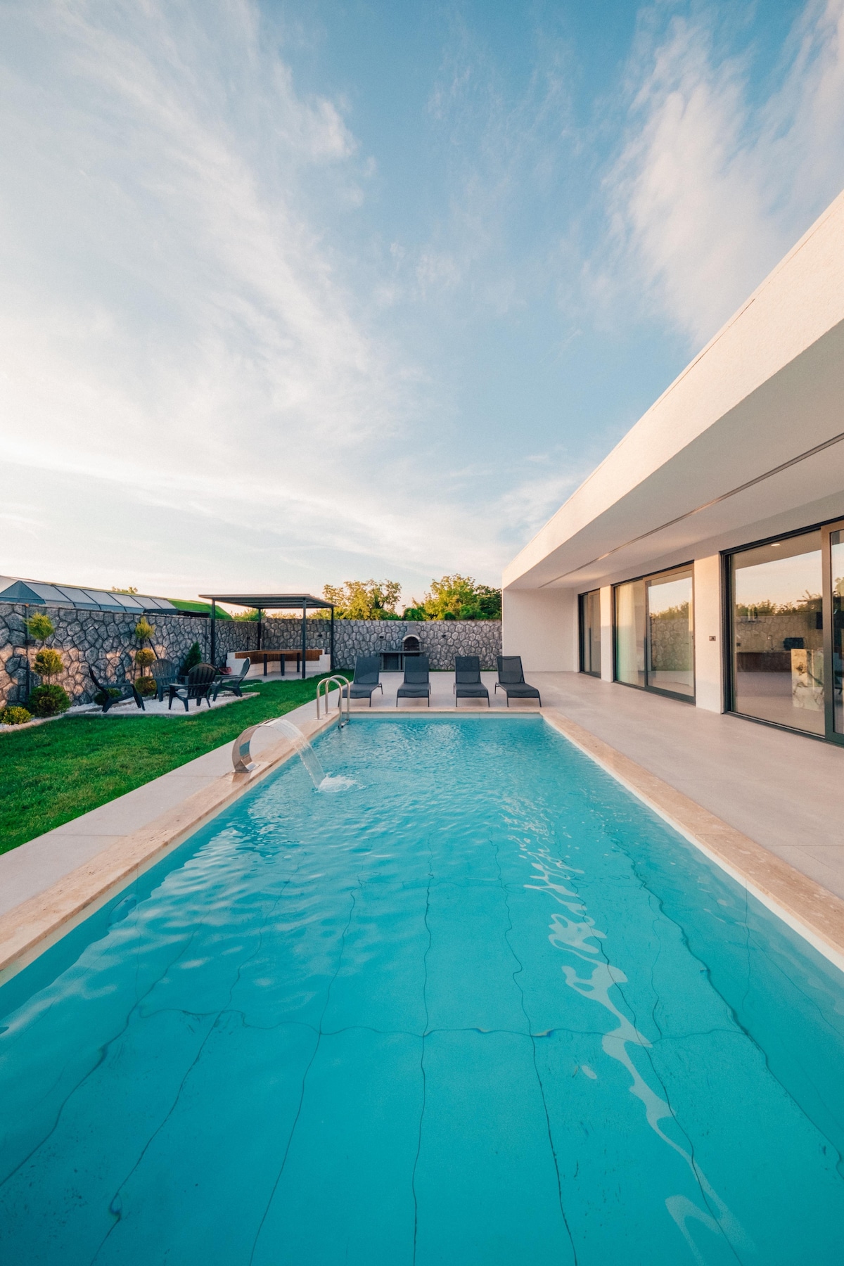 A sleek swimming pool is surrounded by a spacious deck with lounge chairs. Large glass doors offer a view into the bright interior. Lush greenery and a pergola are visible in the background, contributing to a serene outdoor atmosphere.