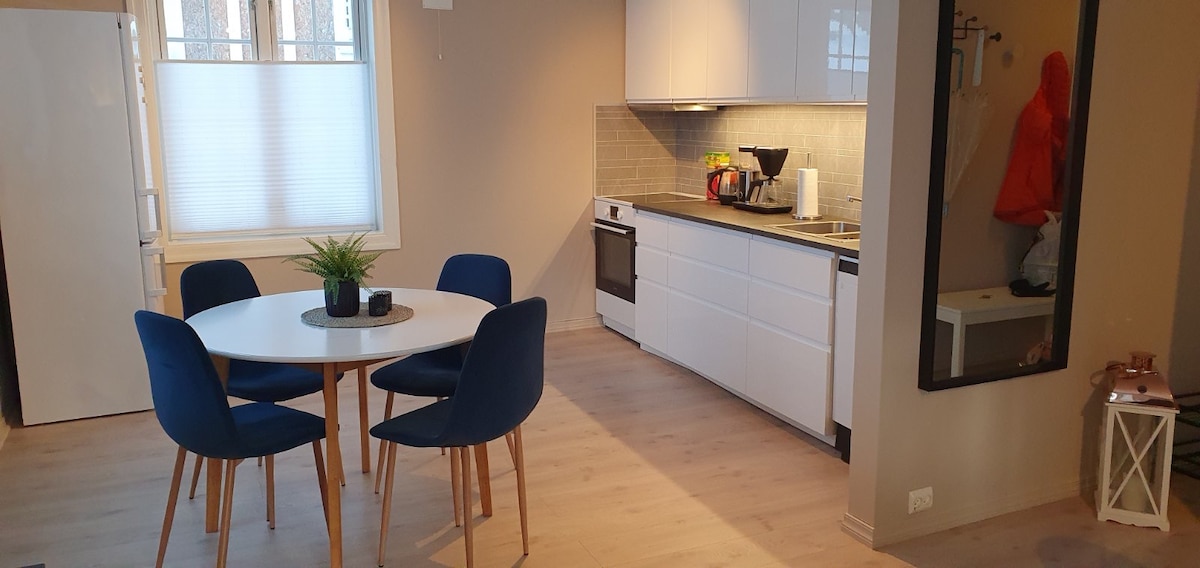A dining area features a round table surrounded by four blue chairs. A kitchen is visible in the background, equipped with white cabinetry and modern appliances. Natural light filters through a window, illuminating the neutral tones of the walls and flooring.