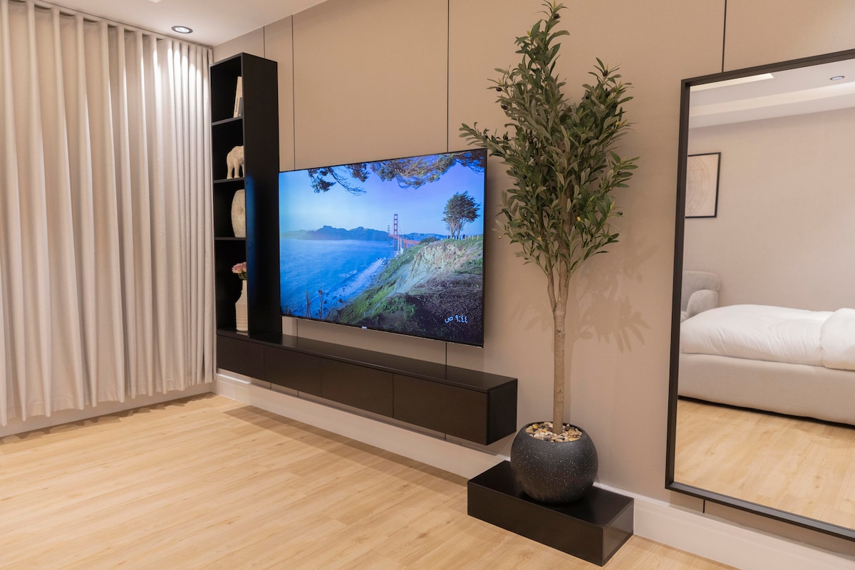 A modern living area is featured with a spacious wooden floor. A large wall-mounted television displays a scenic landscape. A decorative plant adds greenery beside a sleek black console that holds books and ornaments, while a large mirror reflects light from the nearby space.