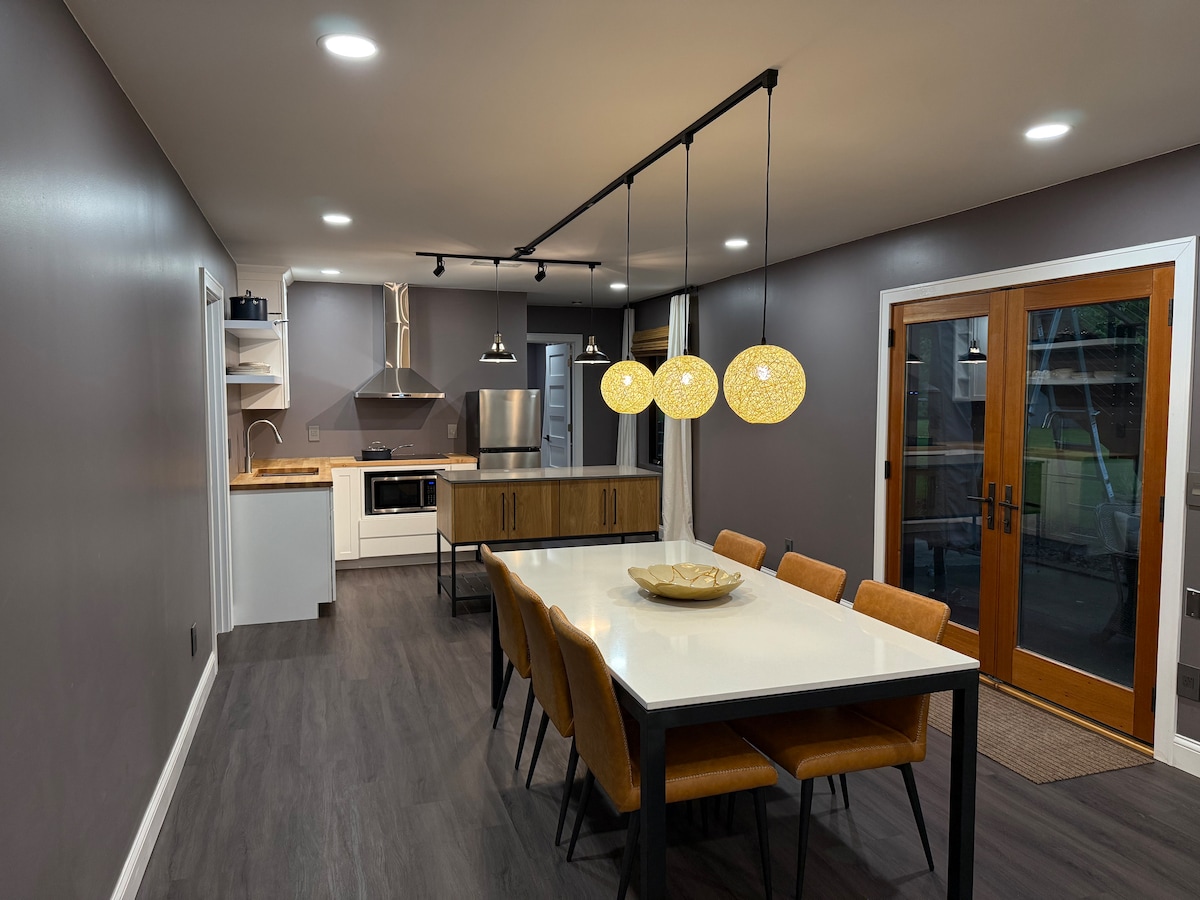A modern kitchen is featured, showcasing a central dining table with six chairs. Pendant lights above provide soft illumination. Appliances include a stainless steel refrigerator and oven. Large glass doors open to an outdoor space, while sleek cabinetry complements the neutral wall tones.