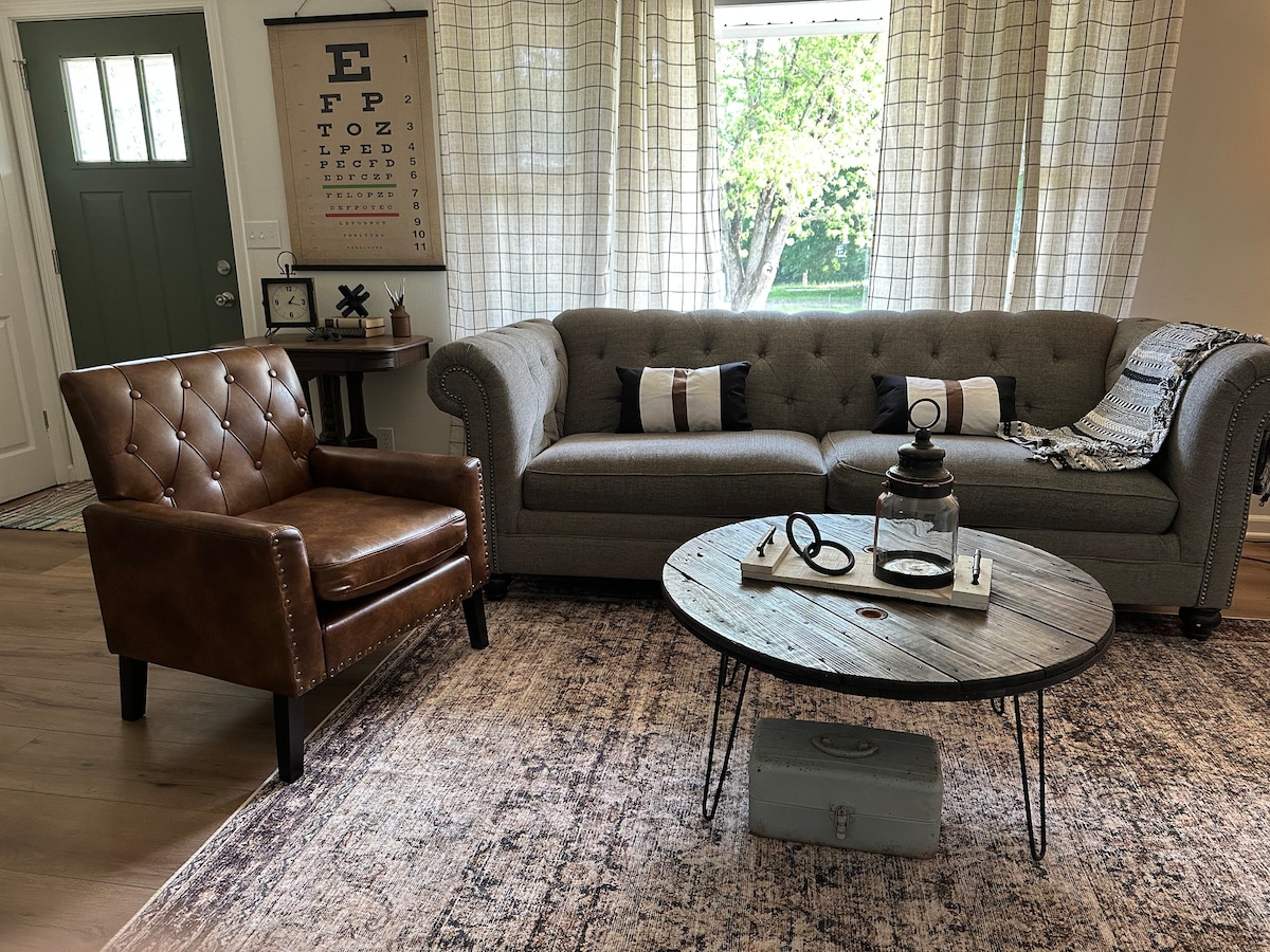 A cozy living area features a tufted armchair alongside a plush sofa, both positioned around a round coffee table. Natural light filters through large windows adorned with light fabric curtains, casting a warm glow on the wood flooring and neutral-toned area rug.