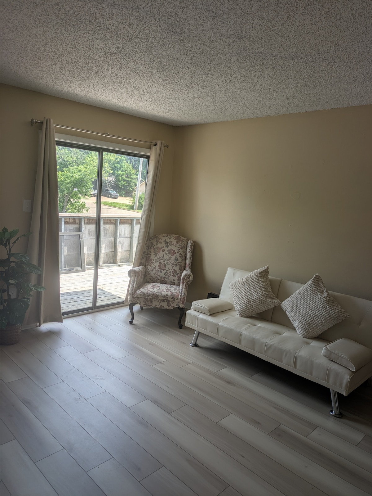 A bright living area features a light-colored sofa bed paired with two decorative cushions. A floral armchair adds charm to the space. Natural light fills the room through sliding glass doors, which open to a wooden deck surrounded by greenery.