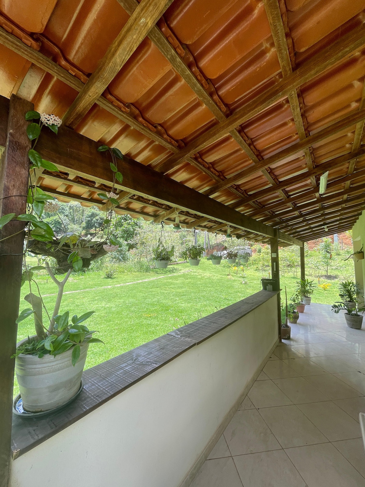 A covered porch features a wooden beam structure and terracotta roof tiles. Potted plants are positioned along the edges, and a lush green lawn extends beyond the porch, showcasing a peaceful garden landscape. Natural light enhances the serene atmosphere.