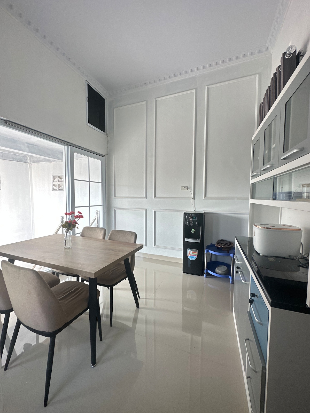 A modern kitchen area is shown, featuring a wooden dining table surrounded by four chairs. Cabinetry lines the wall, complemented by a black countertop hosting a rice cooker. Large glass doors allow natural light to fill the space, while a small floral arrangement adds a touch of elegance.