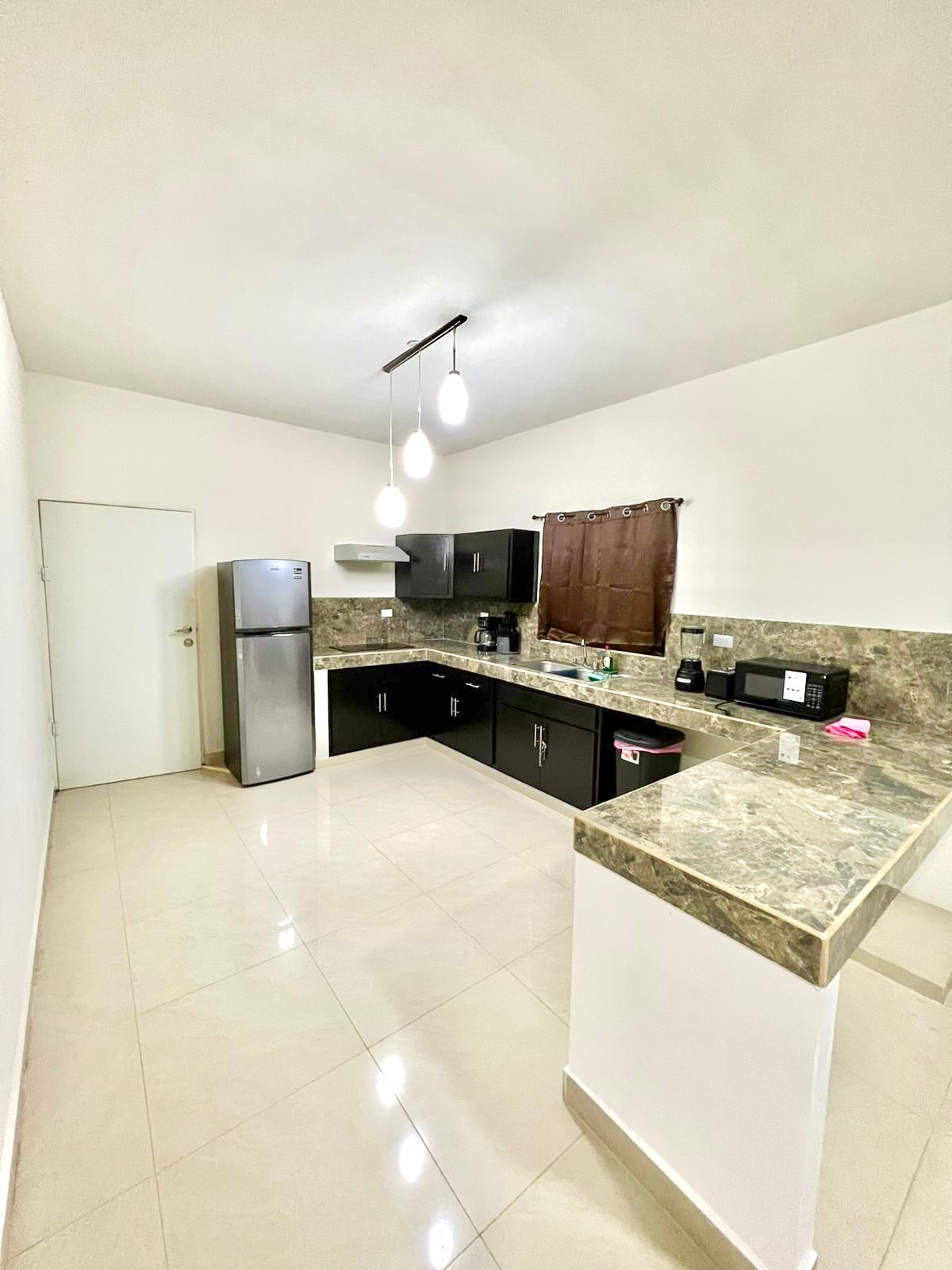 The kitchen features modern appliances including a silver refrigerator, microwave, and black cabinetry, complemented by a stone countertop. The space is well-lit by overhead lights, and a window is covered by a dark curtain, creating an efficient cooking area.