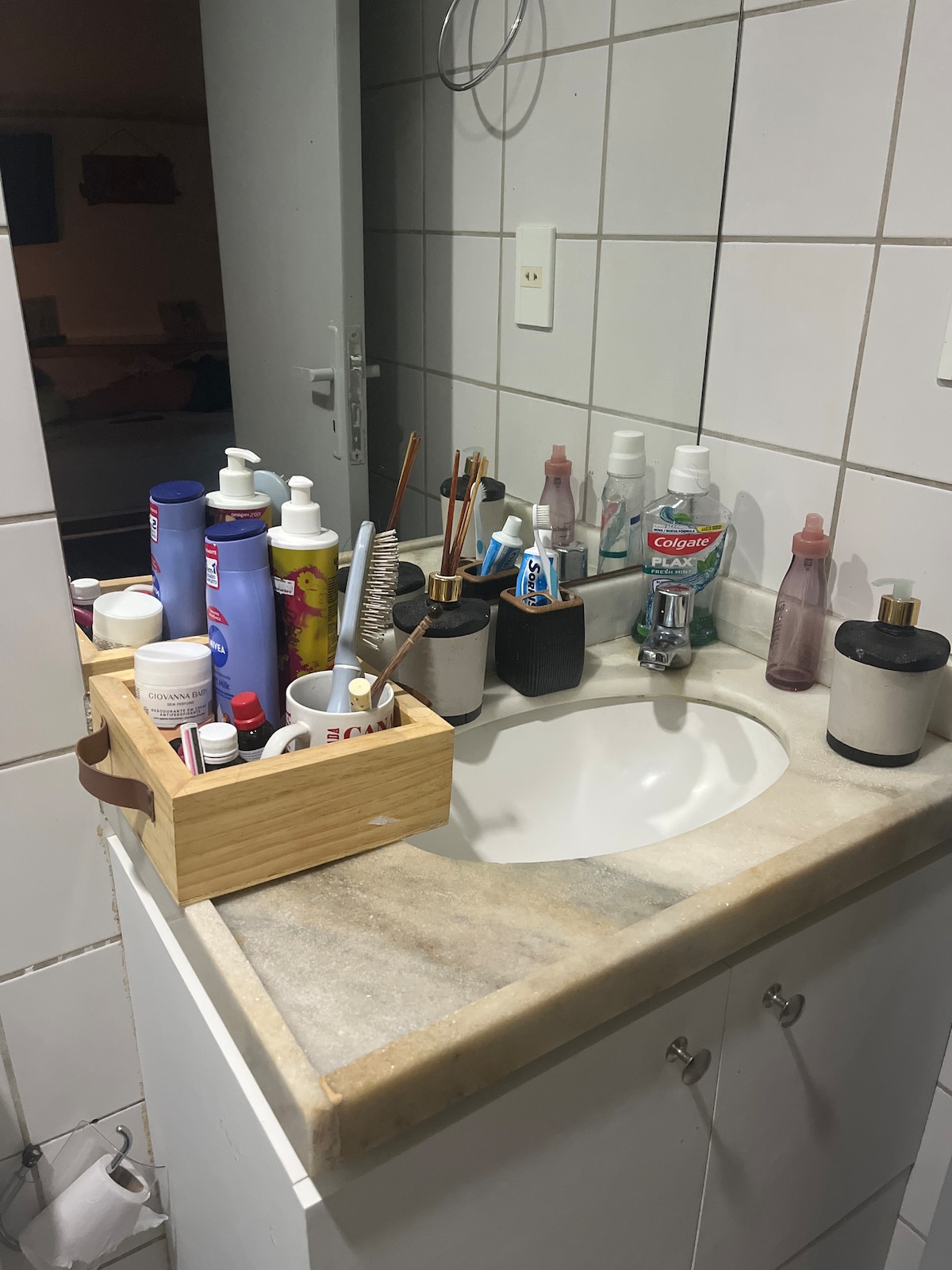 A bathroom countertop is displayed with a range of personal care products, including various bottles and containers. A small wooden tray holds several items while a sink is situated against a tiled wall. The overall arrangement suggests a functional and organized space.