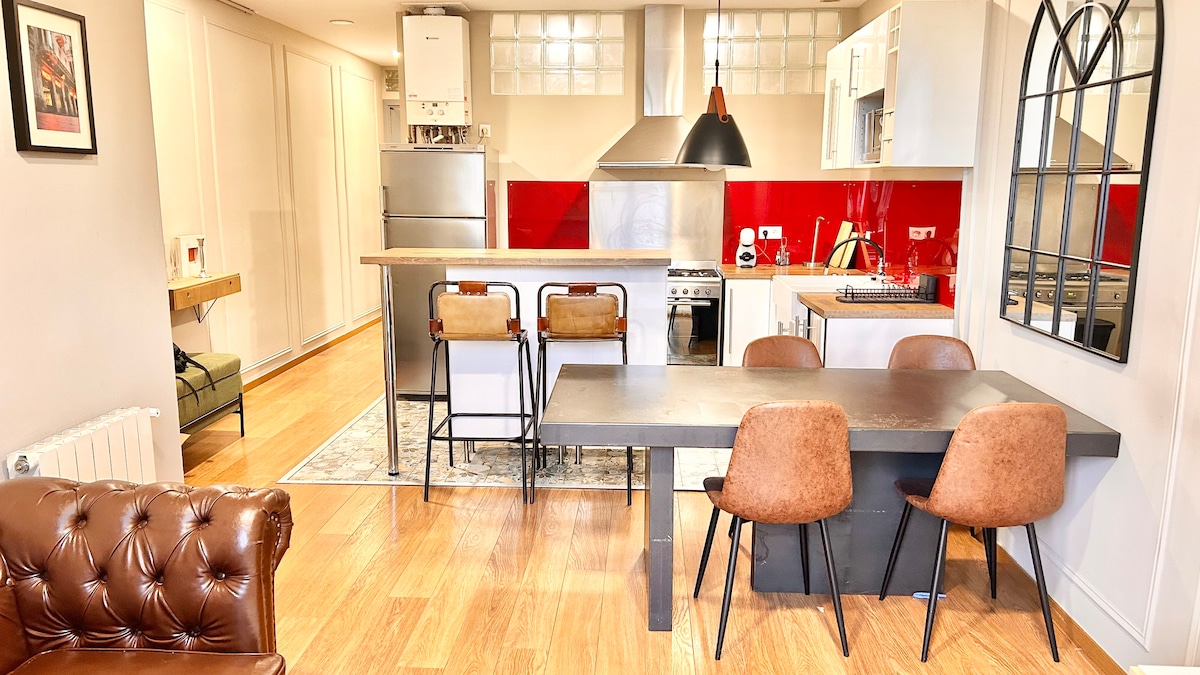 An open-plan kitchen and dining area is displayed, featuring modern stainless steel appliances and red accents. A wooden dining table is surrounded by four upholstered chairs, while two barstools complement the kitchen island. Warm wood flooring enhances the inviting space.