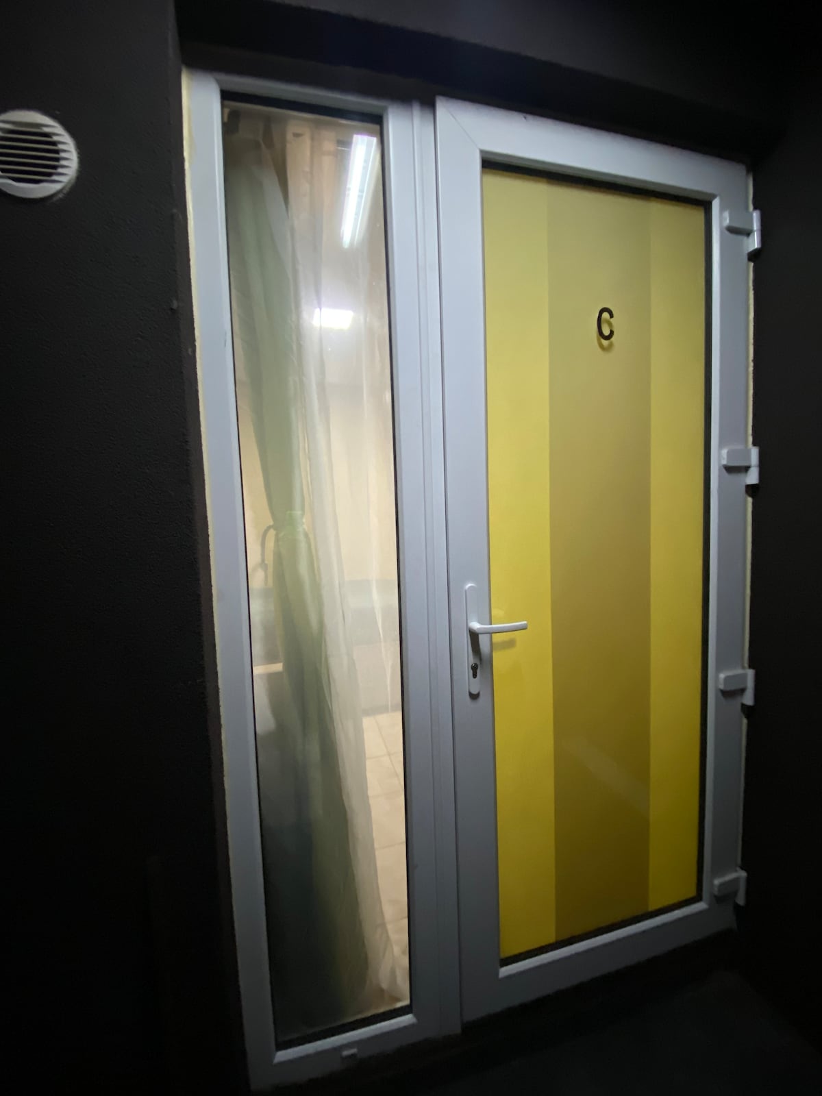A modern exterior door with a yellow panel and a transparent section is visible. The door is framed in white and has a simple handle. Natural light can be seen through the partially covered interior window, reflecting a cozy atmosphere.