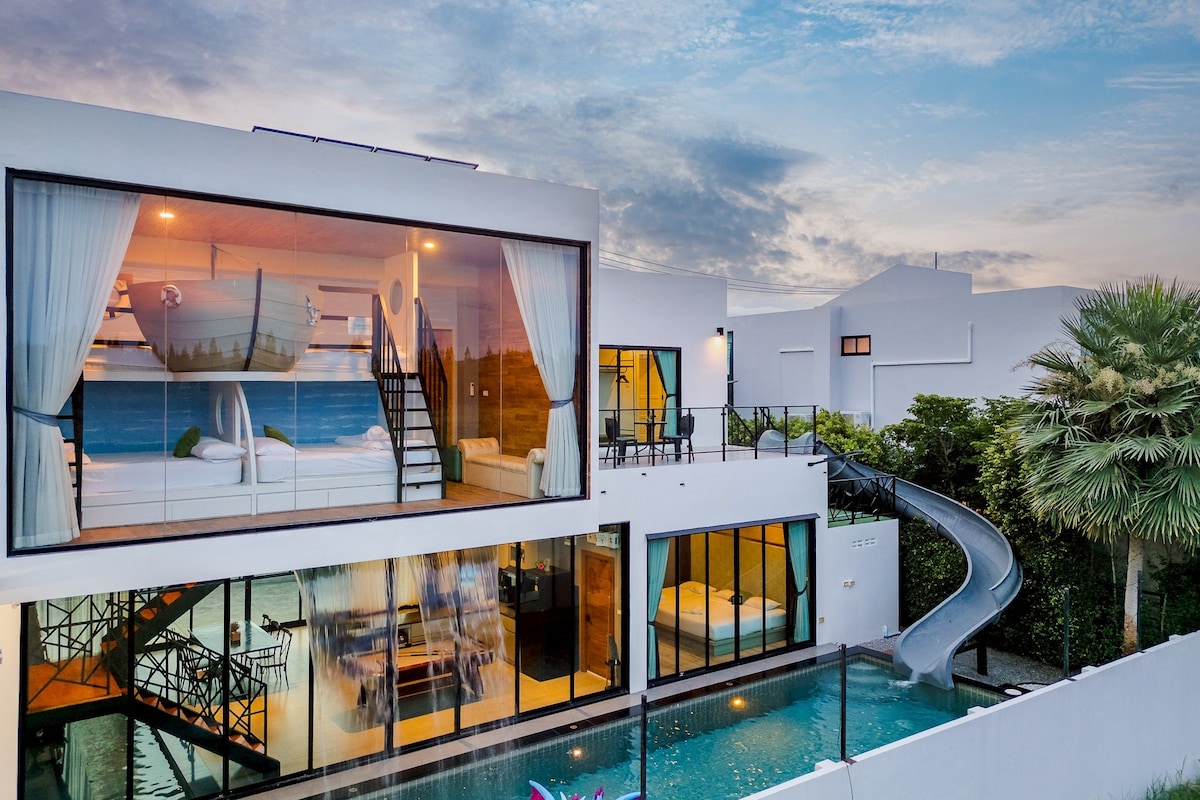 The exterior of a modern villa features large glass windows showcasing the interior layout. A private swimming pool is visible, accompanied by a water slide. Lush greenery surrounds the property, enhancing the tranquil atmosphere as the evening sky transitions in color.