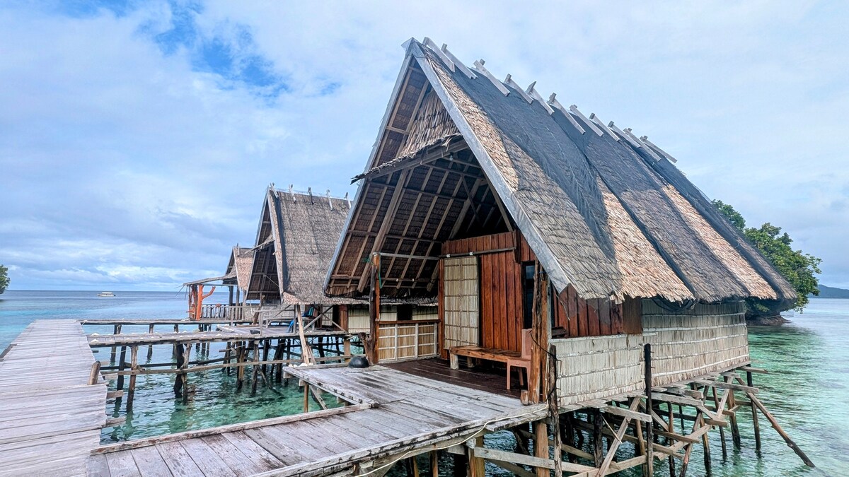 Two traditional bungalows are positioned on stilts over clear turquoise waters. Each bungalow features a thatched roof and wooden exteriors. A wooden walkway connects the bungalows, leading toward the horizon. The surrounding area showcases a serene blend of water and sky.