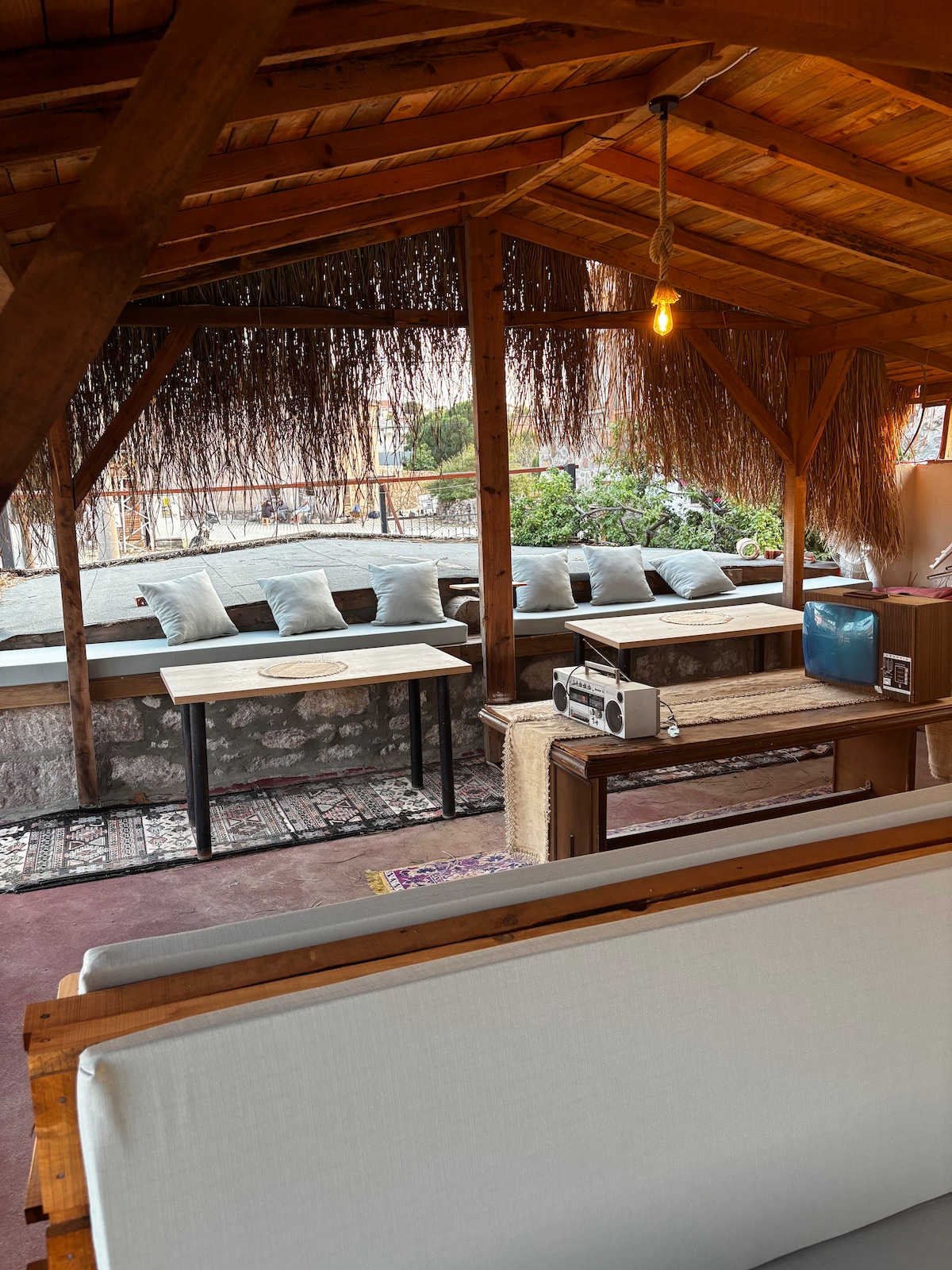 A sunlit seating area showcases a blend of natural wood and comfortable cushions. A long table is placed against the wall, complemented by a retro television and a small audio device. Soft overhead lighting creates a relaxed atmosphere, while greenery is visible through nearby openings.
