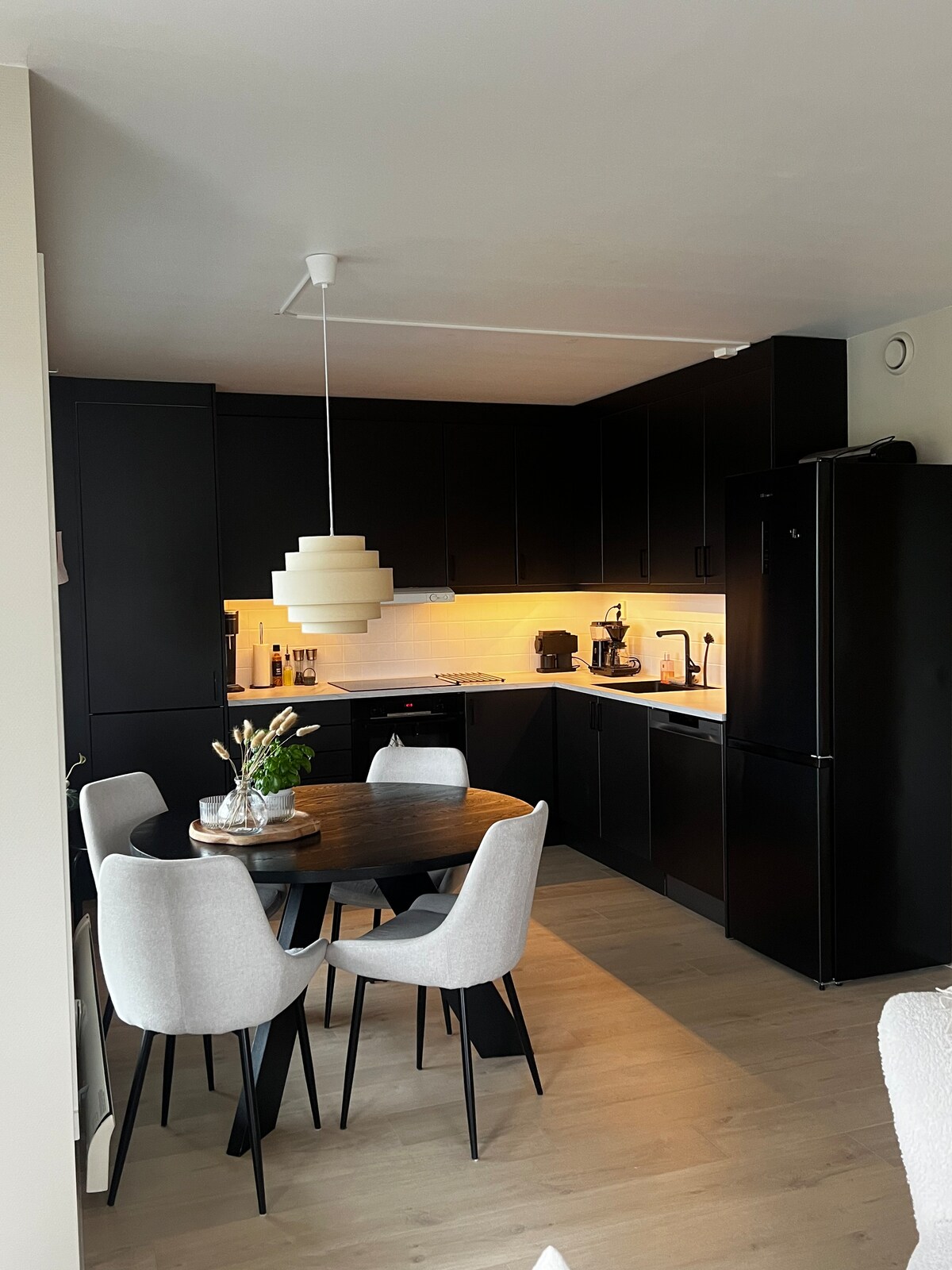 A modern kitchen is presented with sleek black cabinetry and a well-equipped workspace. A round dining table is surrounded by four upholstered chairs. Soft lighting illuminates the countertop, highlighting kitchen appliances, including a coffee maker and refrigerator.