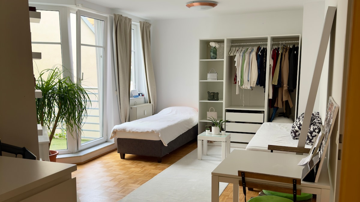 A bright room is featured with large windows allowing natural light to fill the space. A single bed is positioned next to a minimalist white storage unit. A small coffee table sits on a light rug, while a closet holds neatly arranged clothes. Potted plants add a touch of greenery.