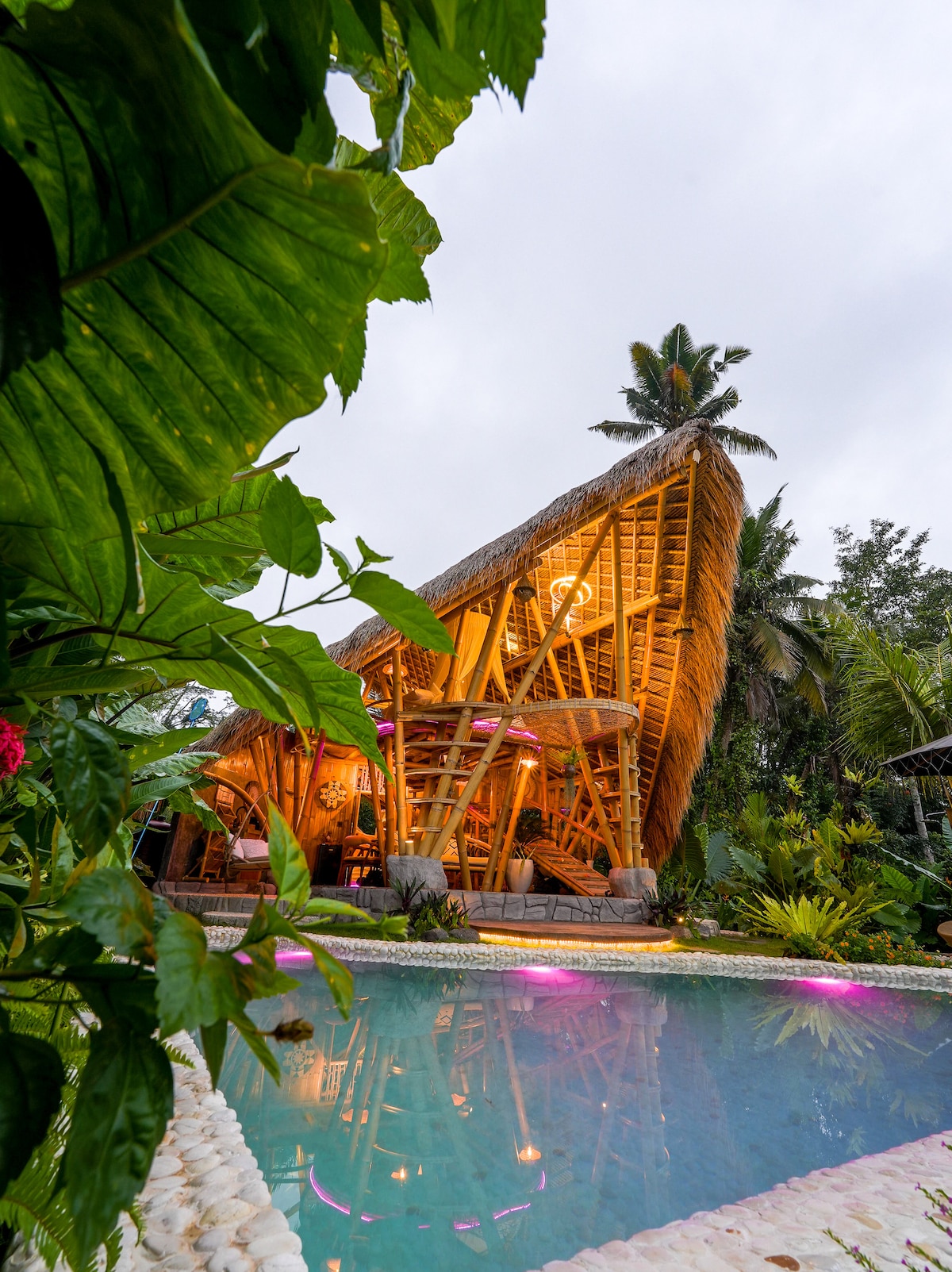 A striking bamboo villa rises gracefully beside a tranquil pool, surrounded by lush greenery. The structure's thatched roof complements the natural setting, while soft lighting creates a warm ambiance. The pool is enhanced with subtle purple lights, inviting relaxation in a serene atmosphere.