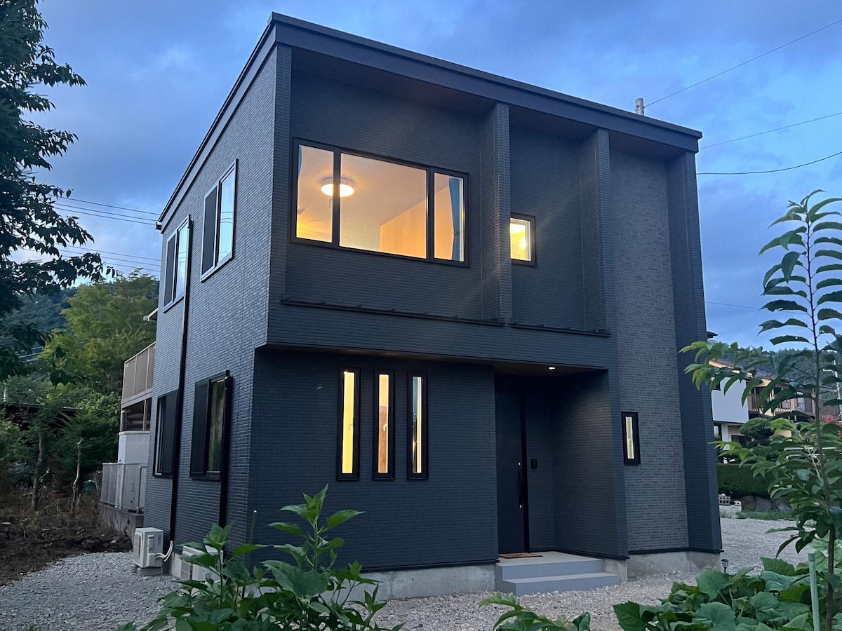 The modern two-story building features a sleek dark exterior, with large windows that highlight the interior's warm lighting. A small garden area surrounds the structure, complemented by a gravel driveway. The setting is enhanced by natural greenery, contributing to the property's inviting presence.