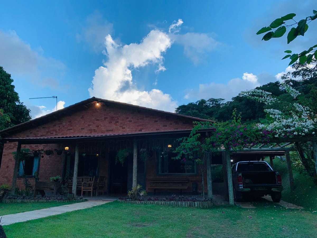 A charming brick house is set in a natural environment, framed by lush greenery and clouds above. A porch area with seating overlooks the well-maintained lawn, and a vehicle is parked under the carport beside the home, enhancing the accessibility of the space.