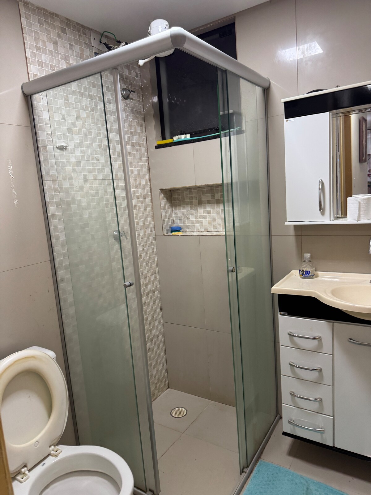A modern bathroom is presented with a glass-enclosed shower featuring neutral tile accents. A white sink with storage cabinets and a mirror reflects the space. Soft blue bath mats are placed on the floor, and a toilet is positioned on the side.