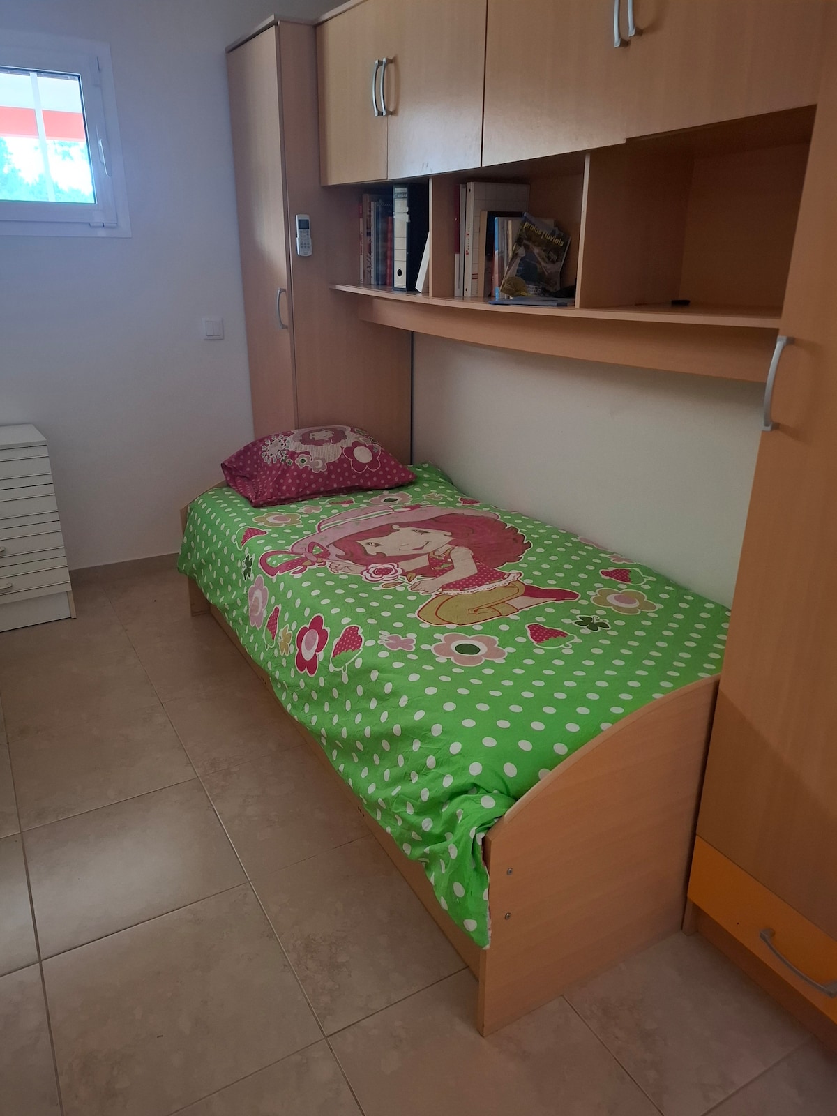 A compact sleeping area features a single bed with a colorful green bedspread adorned with playful patterns. Shelving above holds books and personal items. The room has light tiles on the floor and a window allowing natural light to enter.