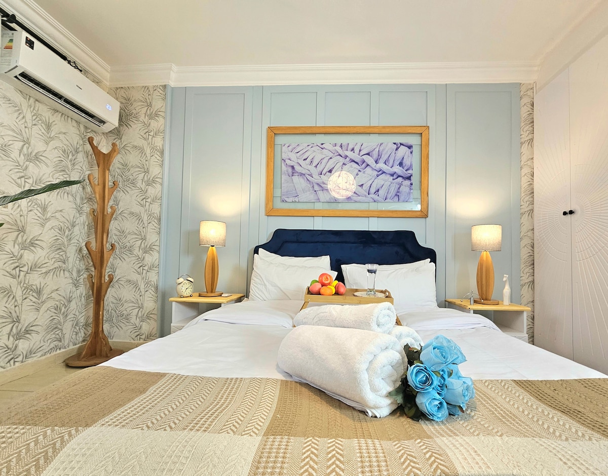 A spacious bed is centered in the room, surrounded by soft wall colors and stylish decor. A piece of art depicting natural forms hangs above. Two lamps provide gentle lighting beside the bed, while a small table displays fresh fruit. Rolled towels and decorative flowers are arranged neatly.