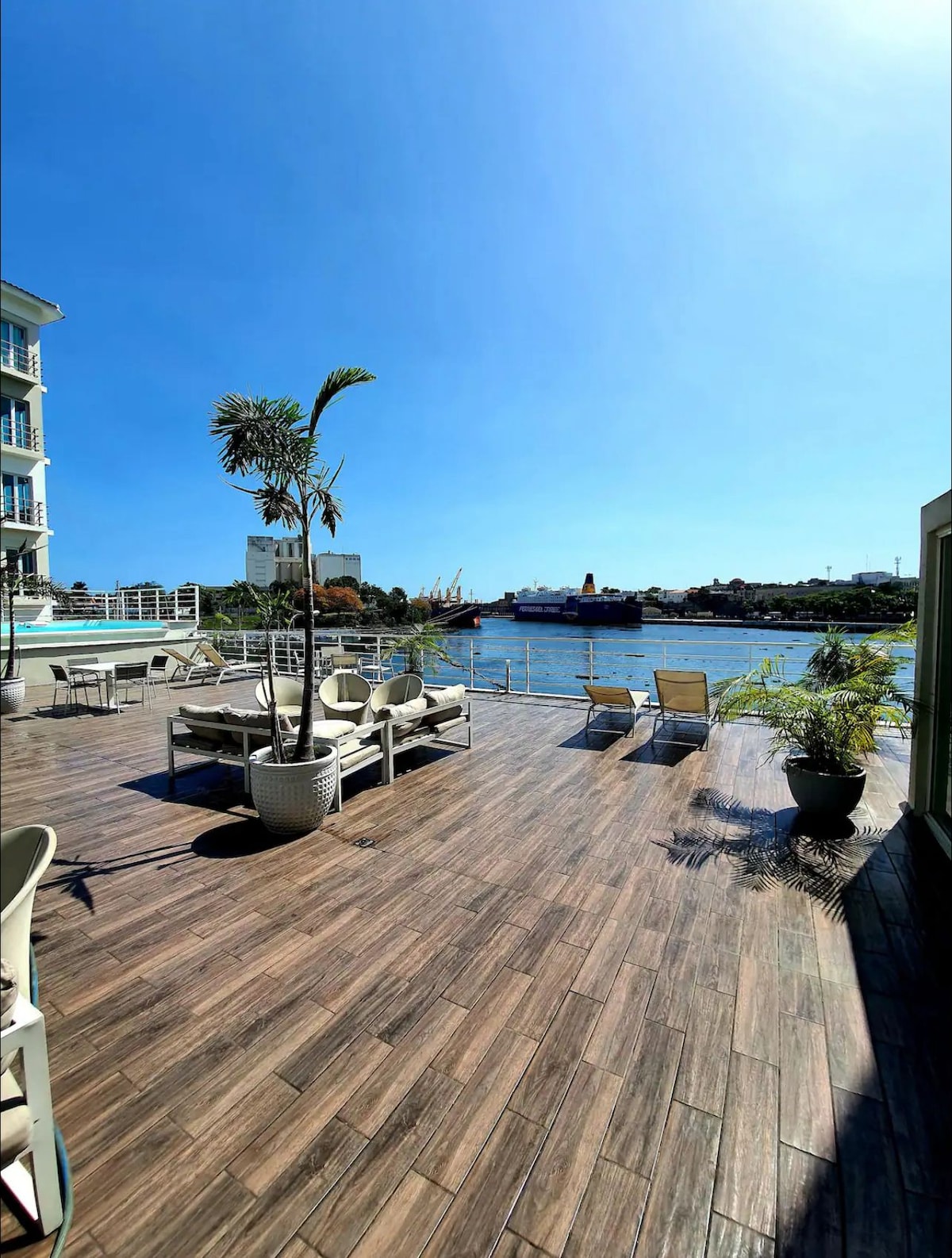 An outdoor terrace features wooden flooring, adorned with lounge chairs and potted plants. The tranquil waters are visible in the background, with a clear blue sky enhancing the serene environment. This space invites relaxation with views of surrounding greenery and the waterfront.