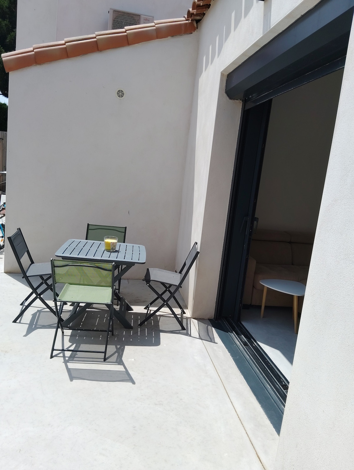 A private terrace is depicted featuring a small dining table paired with four folding chairs. Natural light is visible through an open sliding door, leading into the living area. The smooth floor adds to the outdoor ambiance, providing a relaxed space for guests.