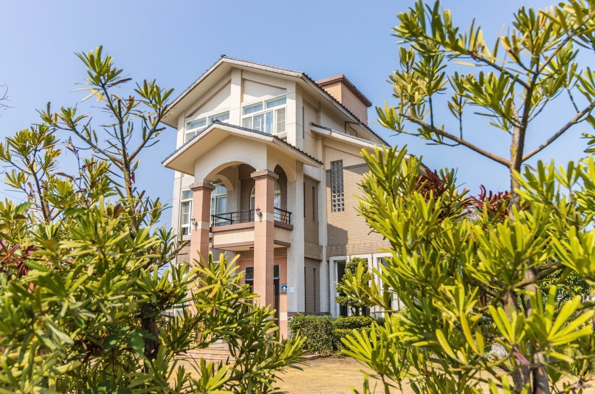 The three-story building is framed by vibrant green foliage, showcasing a blend of beige and white exterior finishes. Architectural features include a prominent arched entrance and large windows, allowing natural light to enhance the living spaces. The clear blue sky provides a serene backdrop.