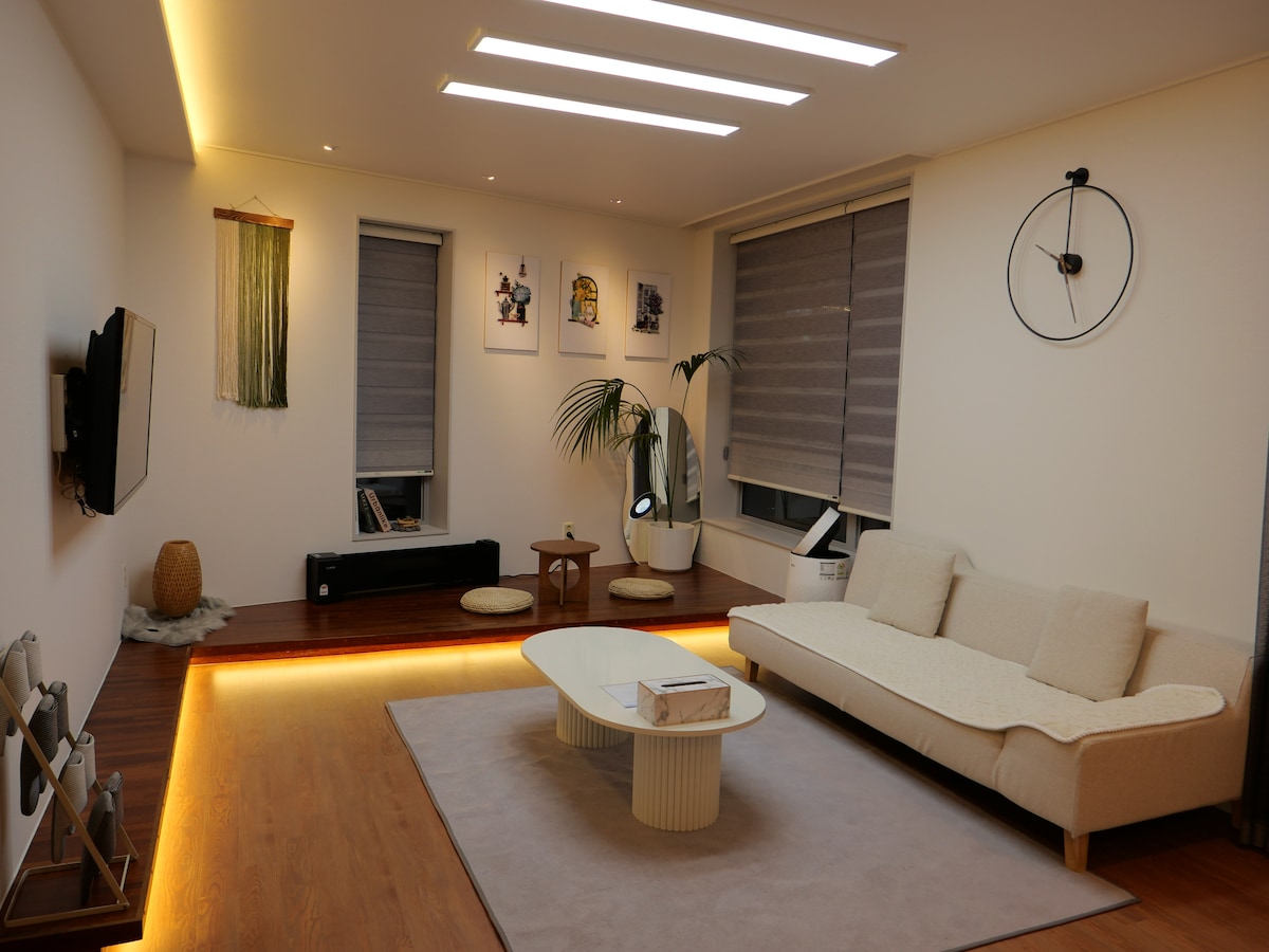 A modern living room features a light-colored sofa and a circular coffee table on a soft area rug. Ambient lighting highlights the wooden floor. Several artworks adorn the walls, and plants add a touch of nature, enhancing the room's spacious feel.