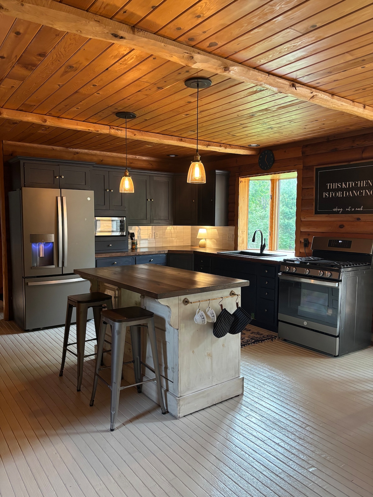 A modern kitchen features dark cabinetry and a central island with seating. Pendant lights illuminate the space, complemented by a large window allowing natural light to fill the room. Stainless steel appliances and a tiled backsplash add a contemporary touch to the rustic surroundings.