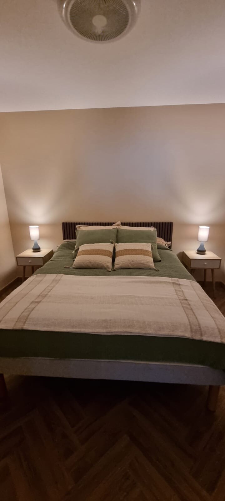 A well-furnished bedroom features a spacious bed with decorative pillows arranged on it. Nightstands with lamps are positioned on either side, providing soft lighting. The walls are painted in a neutral tone, and warm lighting creates a calming environment.