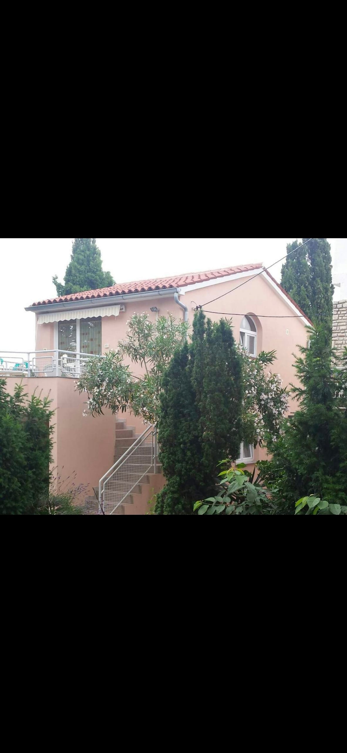 The exterior of the apartment showcases a light-colored building bordered by lush greenery. Staircases lead to the entrance, flanked by tall trees that create a natural ambiance. Large windows allow for light to filter inside, emphasizing a connection with the surrounding environment.