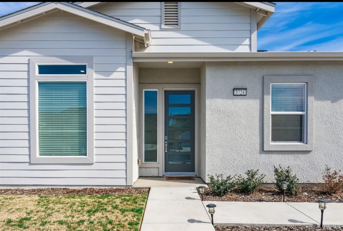 The home's entrance is featured, showcasing a modern front door framed by two large windows on either side. A well-maintained walkway leads to the door, while subtle landscaping elements, such as small shrubs and grass, contribute to the inviting façade.