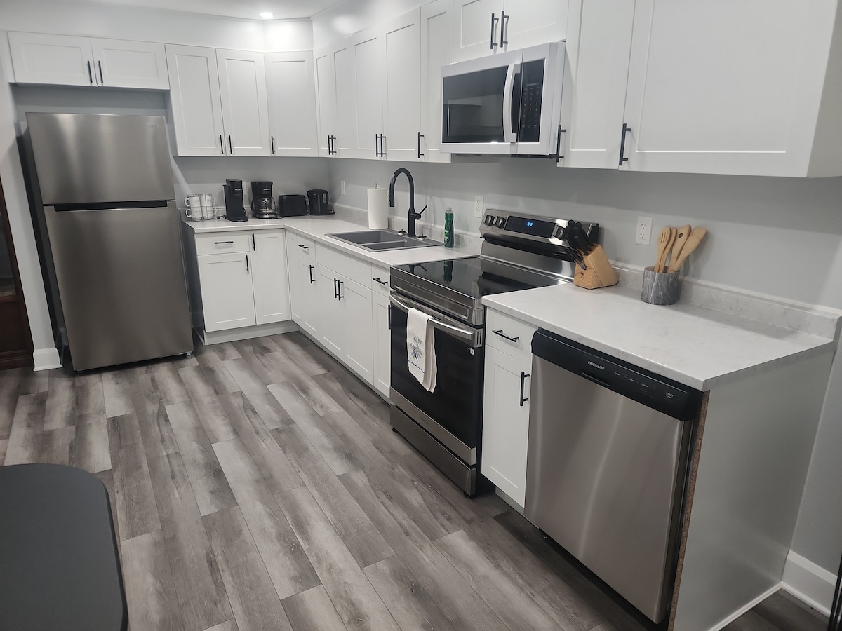 A modern kitchen is equipped with stainless steel appliances, including a refrigerator, microwave, and oven. White cabinetry provides ample storage space, while a large sink sits between the stove and a dishwasher. The countertop is finished in a light color, complementing the wood-look flooring.