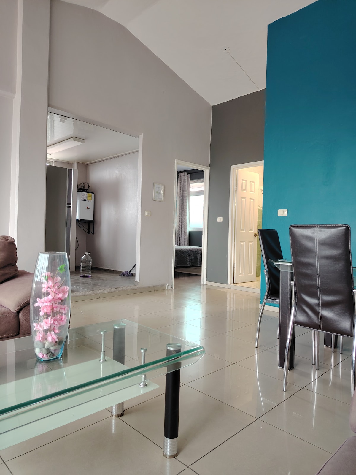 A spacious living area features a low glass coffee table, complemented by dark dining chairs. Light-colored walls are contrasted by a bold blue accent wall. Doorways lead to other areas of the apartment, providing a sense of openness.