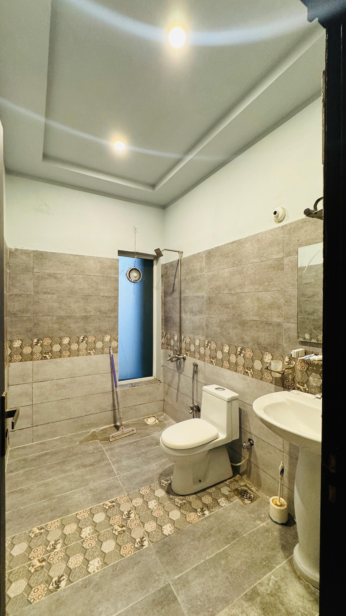 The bathroom features a modern design with a shower area and a toilet. A sink is positioned next to a large mirror. Light-colored tiles with decorative patterns surround the space, and natural light is introduced through a small window.
