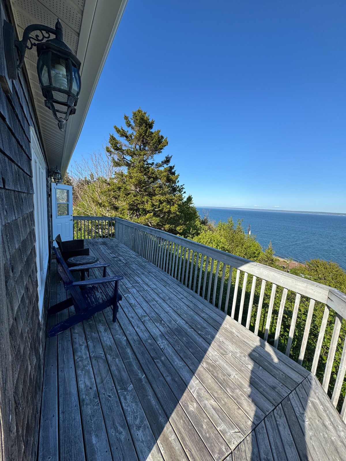 A spacious wooden deck offers views of the ocean and surrounding greenery. Two dark chairs are positioned for relaxation, while a lantern hangs nearby. The sky is clear and blue, enhancing the serene coastal ambiance.