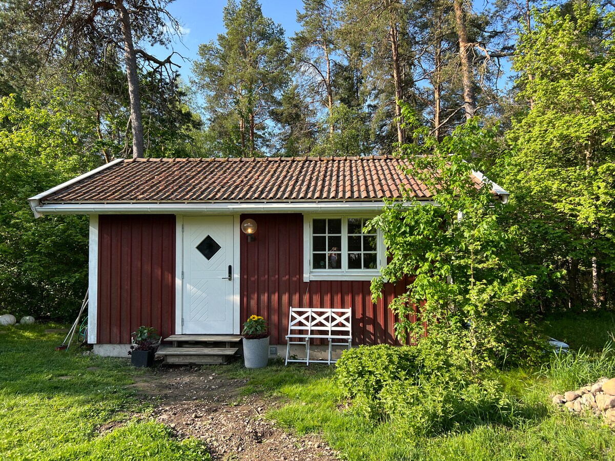 A small red cottage with a tiled roof is surrounded by greenery. The entrance features a white door with a diamond-shaped window and a lantern beside it. A white bench sits in front, and flowerpots add color to the pathway leading to the door.