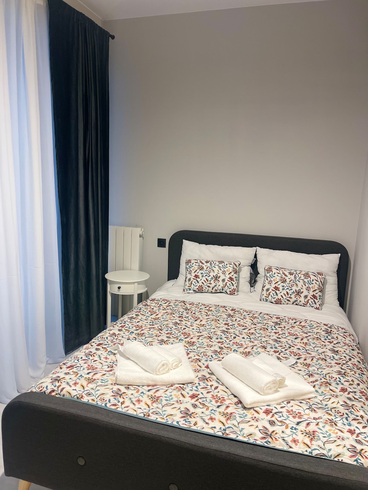 A neatly arranged sleeping area features a double bed adorned with a floral-patterned coverlet and crisp white linens. Two decorative pillows sit at the head of the bed. Soft light filters through sheer curtains, illuminating the space, which includes a small side table.