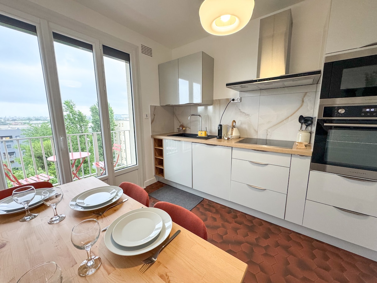 A modern kitchen features sleek cabinetry and a countertop with an integrated sink. A dining table is set with plates and glassware for four, while large windows provide natural light and a view of the surrounding area. A balcony is visible beyond.