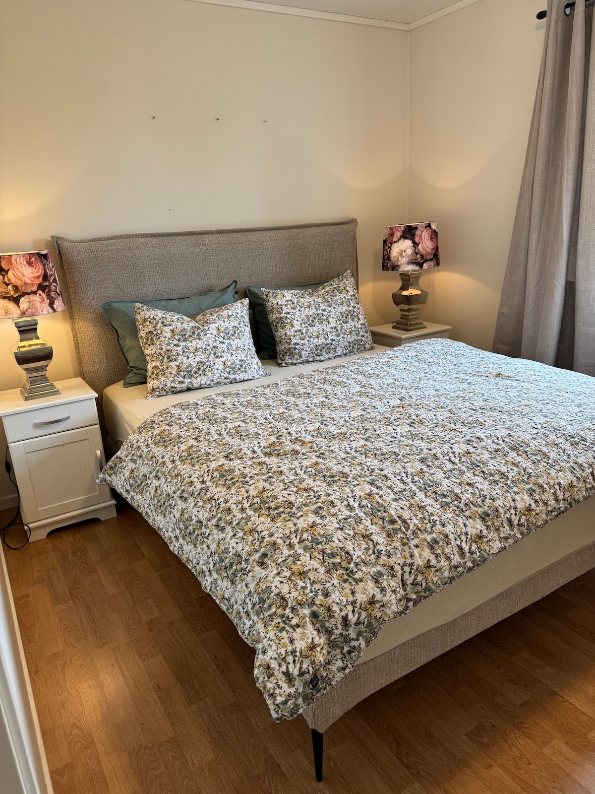 A cozy bedroom features a spacious bed dressed with a floral-patterned coverlet and matching pillows. Adjacent nightstands hold softly glowing lamps. Natural light filters in through a window draped with gray curtains, creating a calm and inviting ambiance.