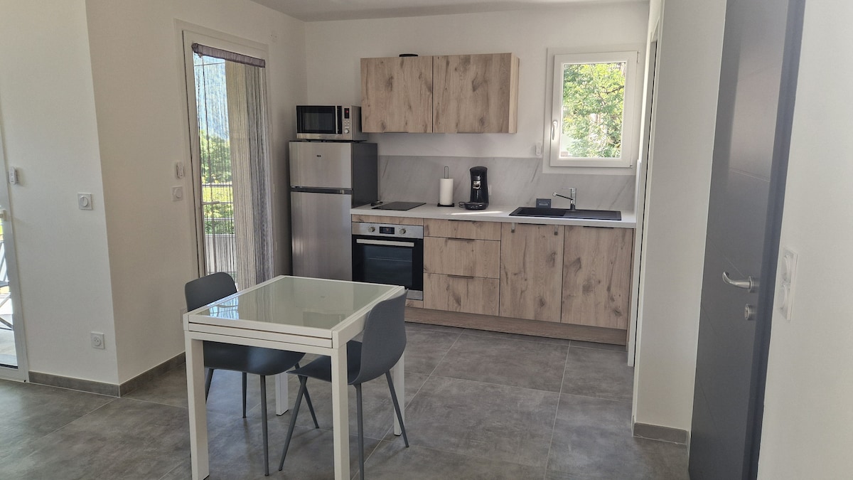 The kitchen area features modern wooden cabinetry and a well-integrated dining space with a glass table and two gray chairs. Appliances include a refrigerator, microwave, and oven. Natural light enters through a window, illuminating the sleek countertops and minimalist design.