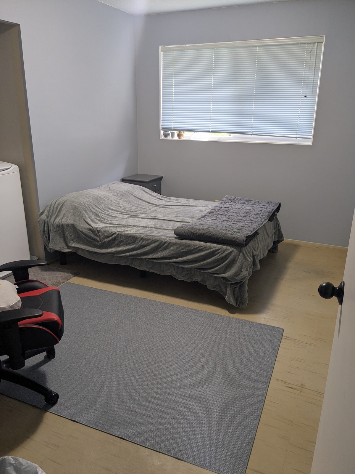 A simple bedroom features a spacious bed, covered with a gray blanket, and a dark bedspread laid across the top. A small nightstand is positioned beside the bed, and a window with blinds allows natural light to enter. A chair and a rug are present on the floor.